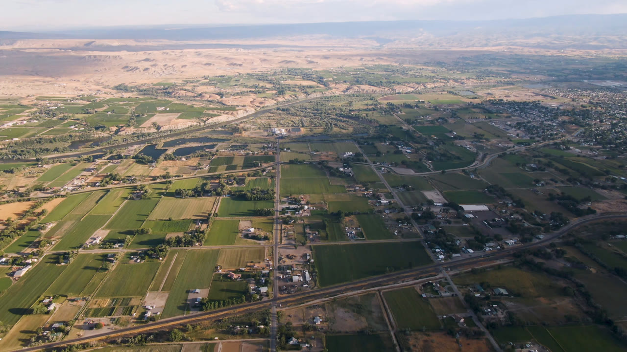 Aerial frame over Palisades, Colorado, with rectangular agricultural fields, a visible highway crossing the lower view, clustered buildings, distant dry hills, and no visible people.