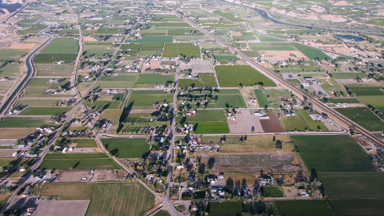 Aerial frame over Palisades, Colorado with a diagonal road, patchwork fields, small building clusters, a river area in the distance, pale hills, and no visible people.