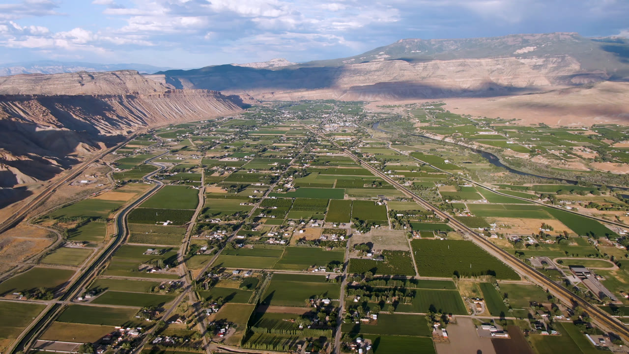 Wide aerial view of green fields, roads, cliffs, and distant mountains under clouds.