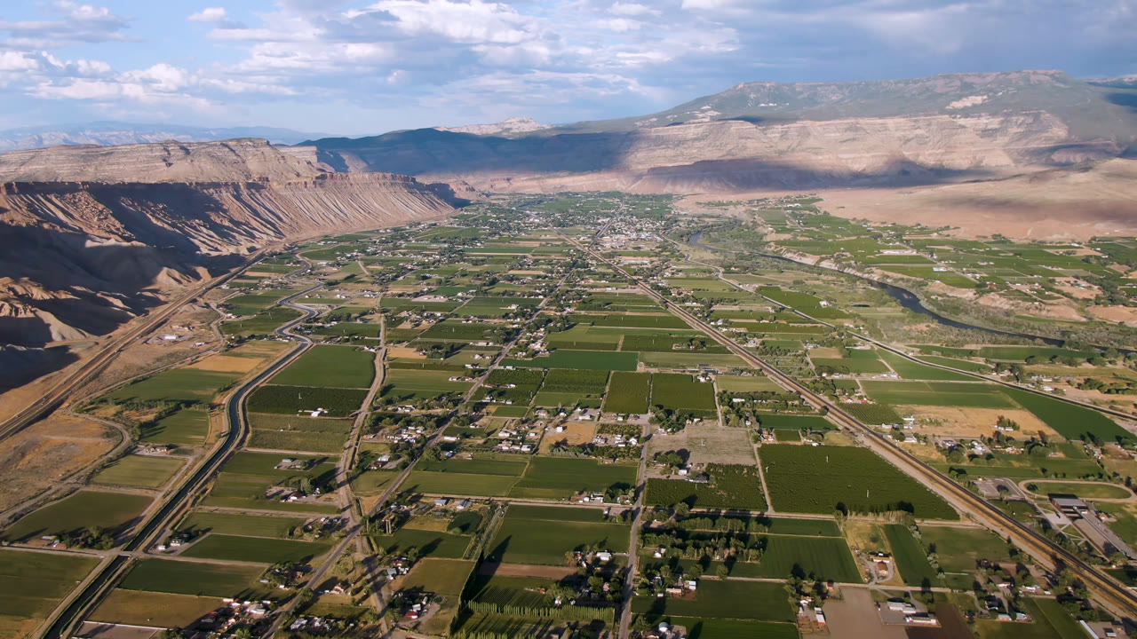 Later aerial frame over Palisades, Colorado with the road corridor more centered, rectangular green fields below, town blocks in the distance, mesas behind them, and no visible people.