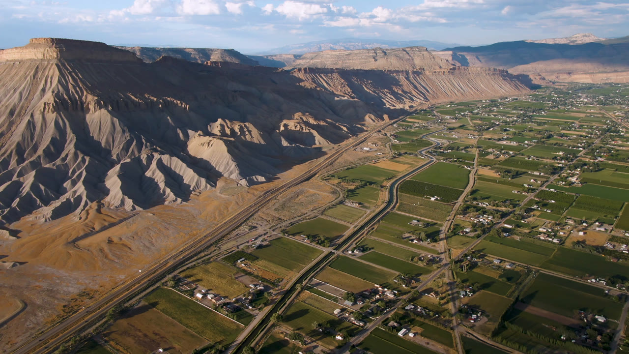 Opening aerial frame over Palisades, Colorado with green farm plots, scattered houses, road corridors, tan canyon cliffs, distant mesas, and no visible people.