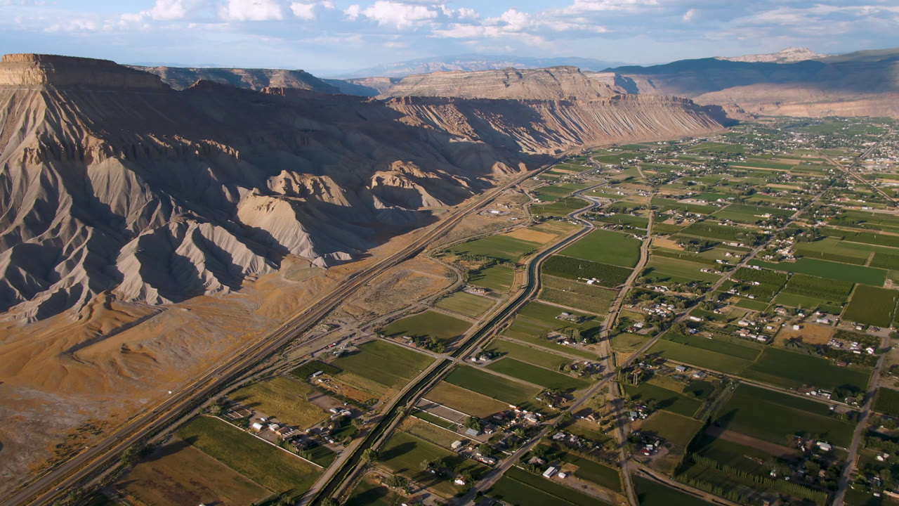 Aerial view of canyon cliffs, green valley fields, roads, and scattered buildings under blue sky.