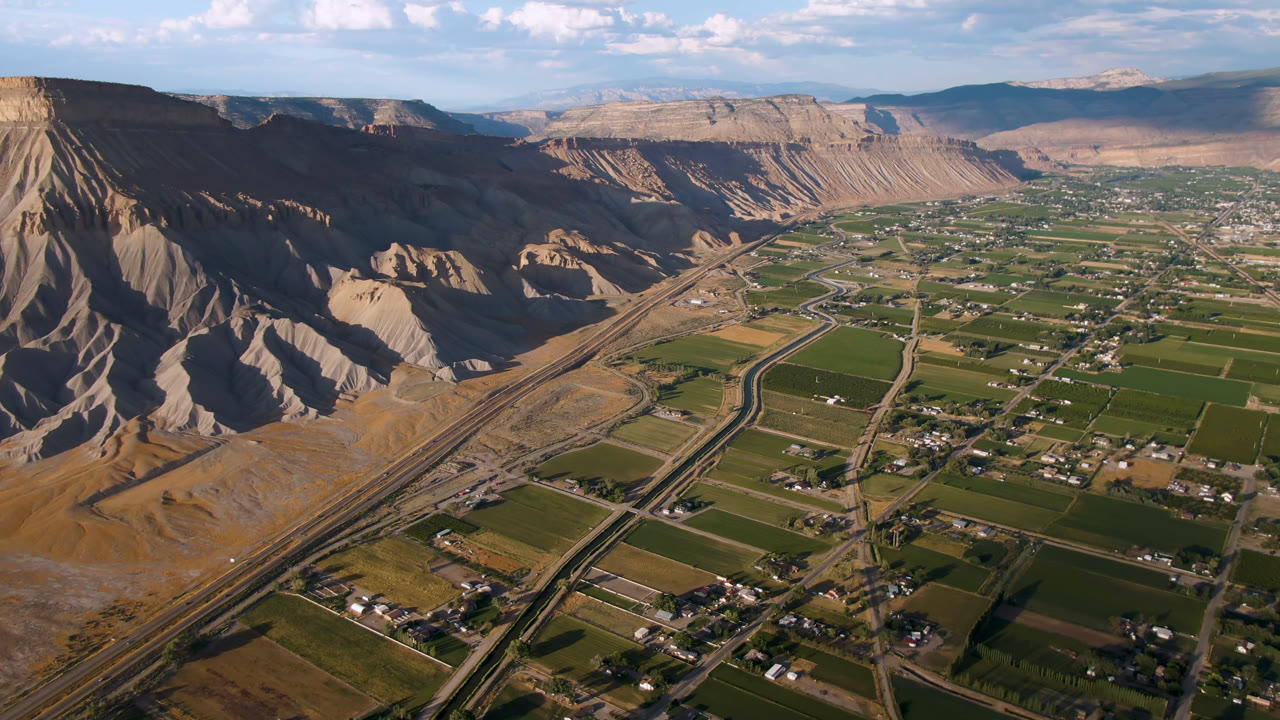 Second aerial frame in Palisades, Colorado, showing more of the valley floor with road and rail-like corridors, patchwork fields, small buildings, canyon slopes, and no visible people.
