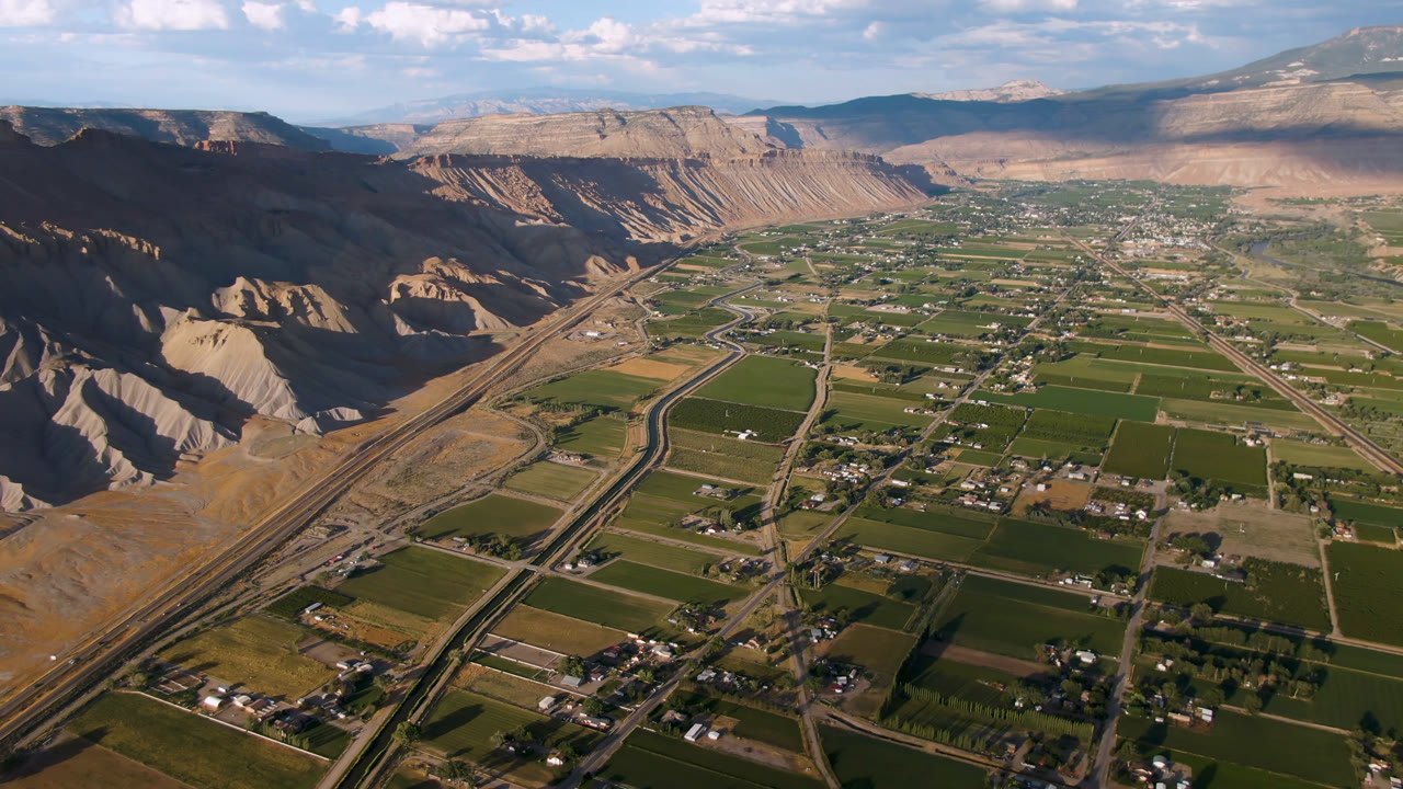Aerial view over Palisades, Colorado, showing green agricultural fields, roads, scattered buildings, dry canyon-like slopes, distant mesas, blue sky, and no visible people.