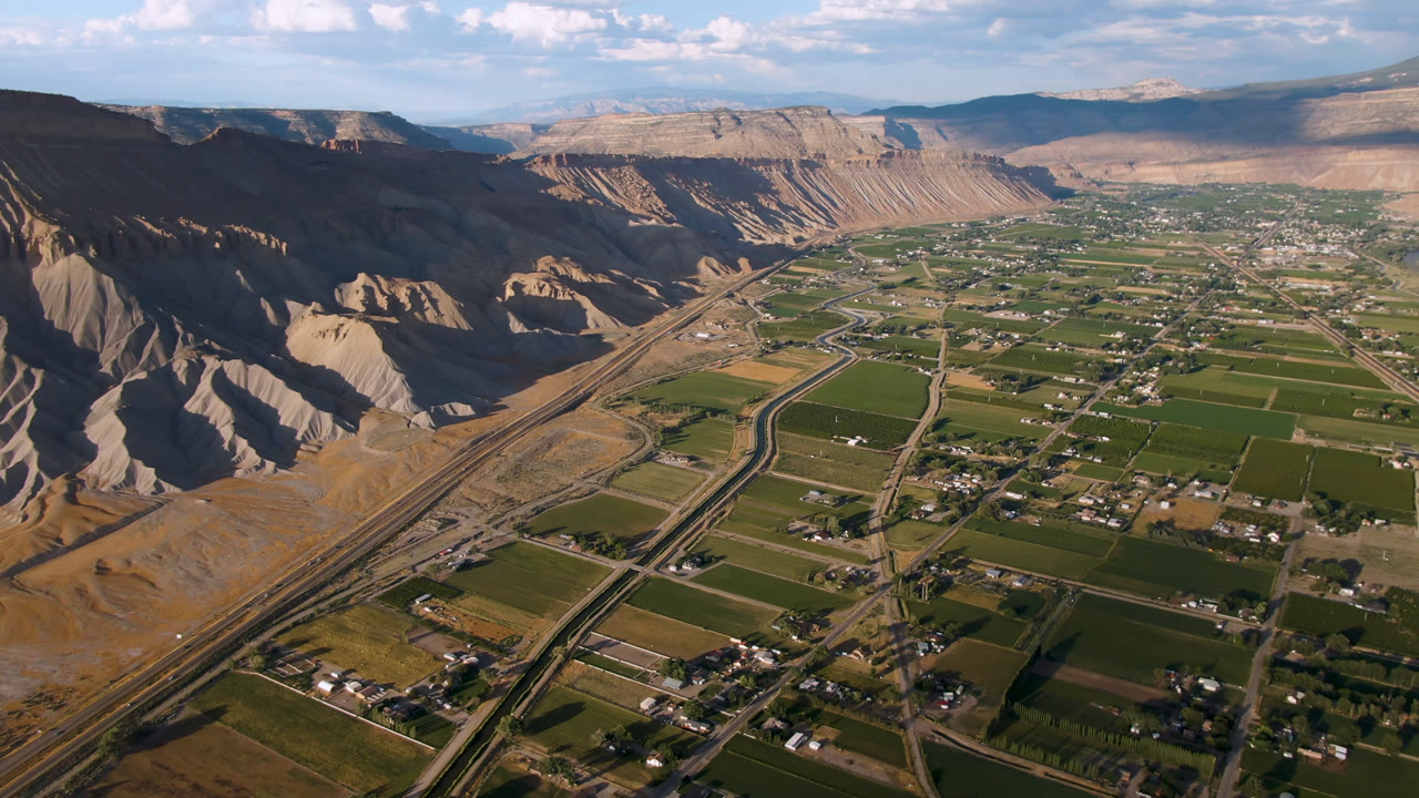 Aerial view of green farm fields, roads, homes, dry ridges, and distant mountains under a blue sky.