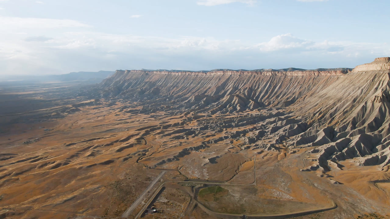 Wide aerial frame over Palisades, Colorado, showing a long eroded mesa, dry flats, a road crossing the lower foreground, distant haze, and no visible people.