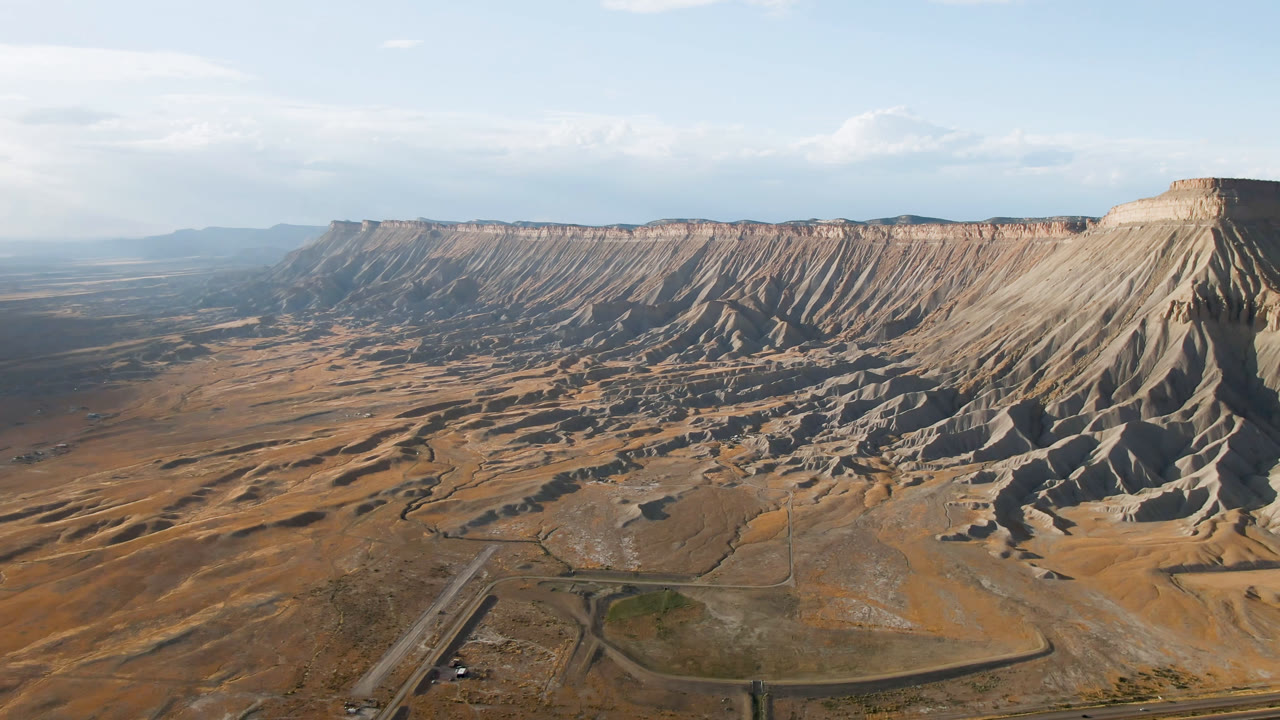Wide aerial frame of Palisades, Colorado showing a tan mesa ridge, eroded slopes, foreground road lines, pale blue sky, and no visible people.