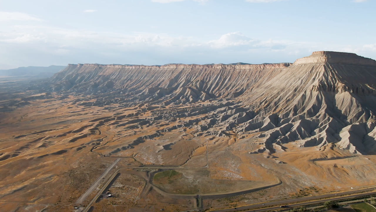 Aerial frame from Palisades, Colorado showing a broad mesa wall, eroded tan slopes, foreground roads, dry flats, and no visible people.