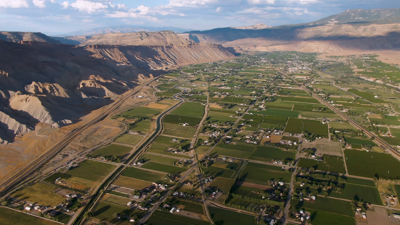 Aerial view over Palisades, Colorado showing green farm fields, scattered homes, roads, rail lines, tan cliffs, distant mountains, scattered clouds, and no visible people.