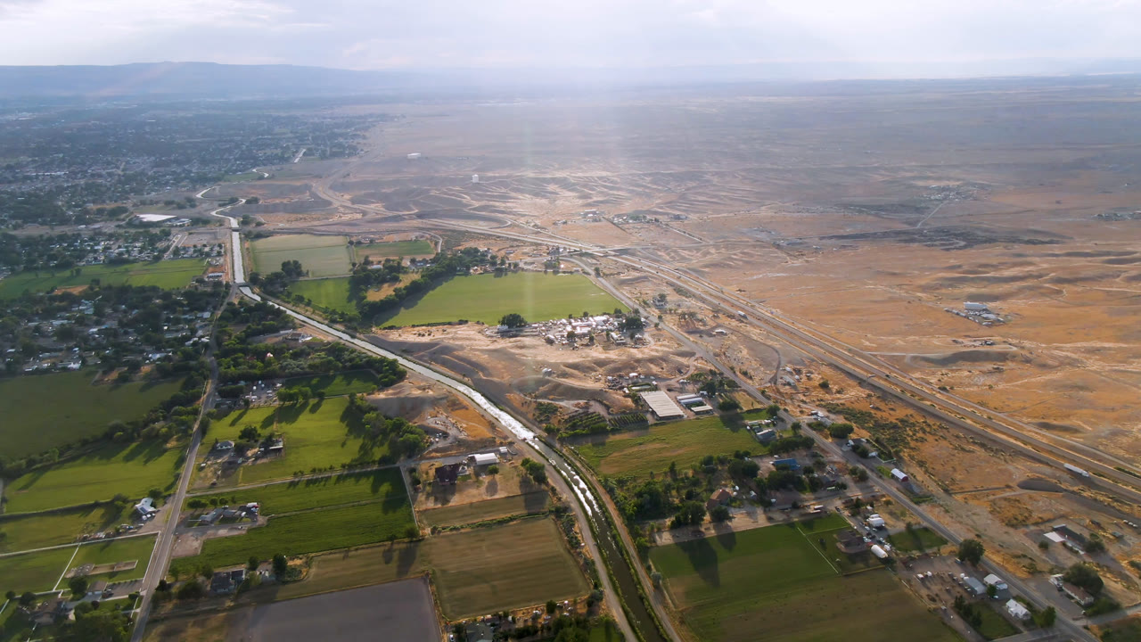 High aerial frame over Palisades, Colorado, with green fields in the lower left, a road corridor through scattered buildings, tan open land beyond, distant hills, and no visible people.