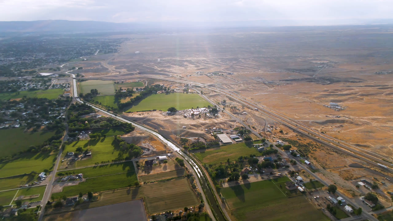 Aerial view of Palisades, Colorado, with a bright green field near the center-left, a winding road entering the rural area, highway lines crossing dry terrain, and no visible people.