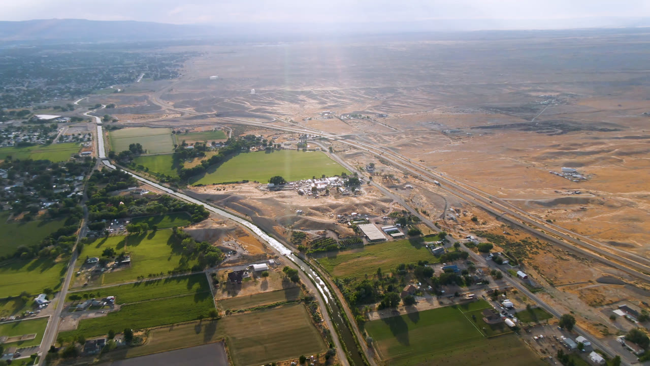 Wide aerial view of rural roads, fields, buildings, and open landscape under hazy sky.