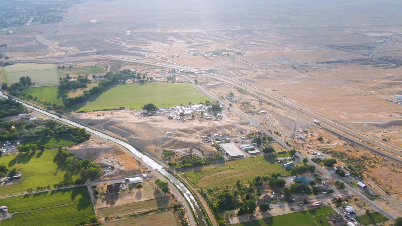 A high aerial view over Palisades, Colorado shows green fields, roadside trees, scattered buildings, dry tan terrain, and no visible people at the start of the clip.