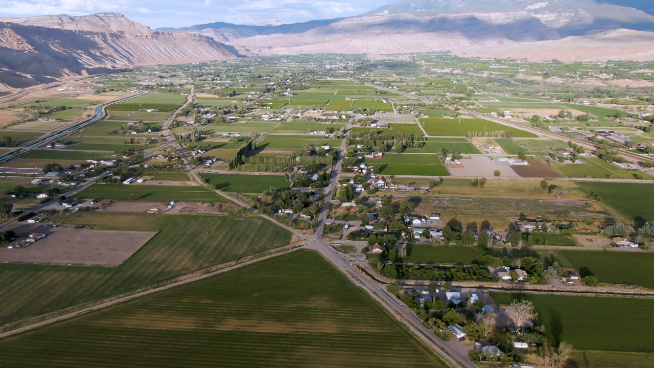 Opening aerial frame over Palisades, Colorado shows rectangular farm fields, a road junction, scattered homes, distant mountains, blue sky, and no visible people.