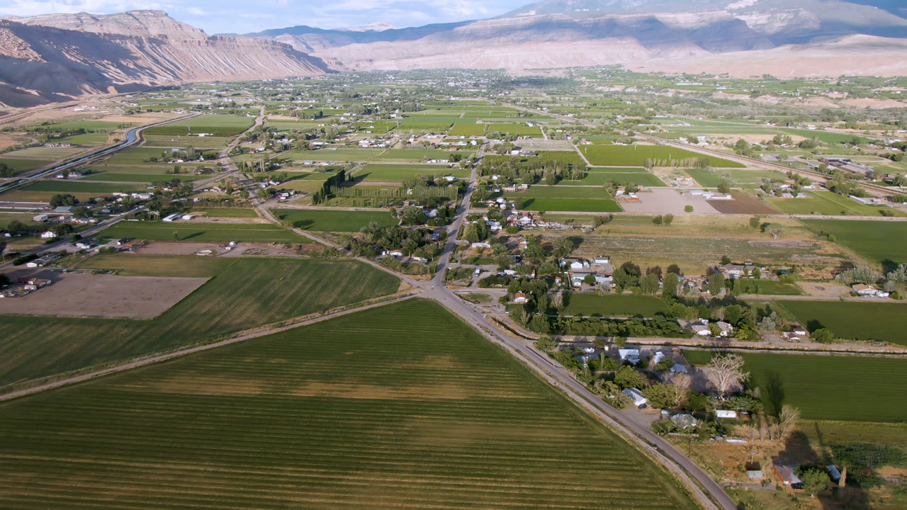 Early aerial frame of Palisades, Colorado showing foreground crop rows, a central road network, scattered homes, pale foothills, distant mountains, and no visible people.