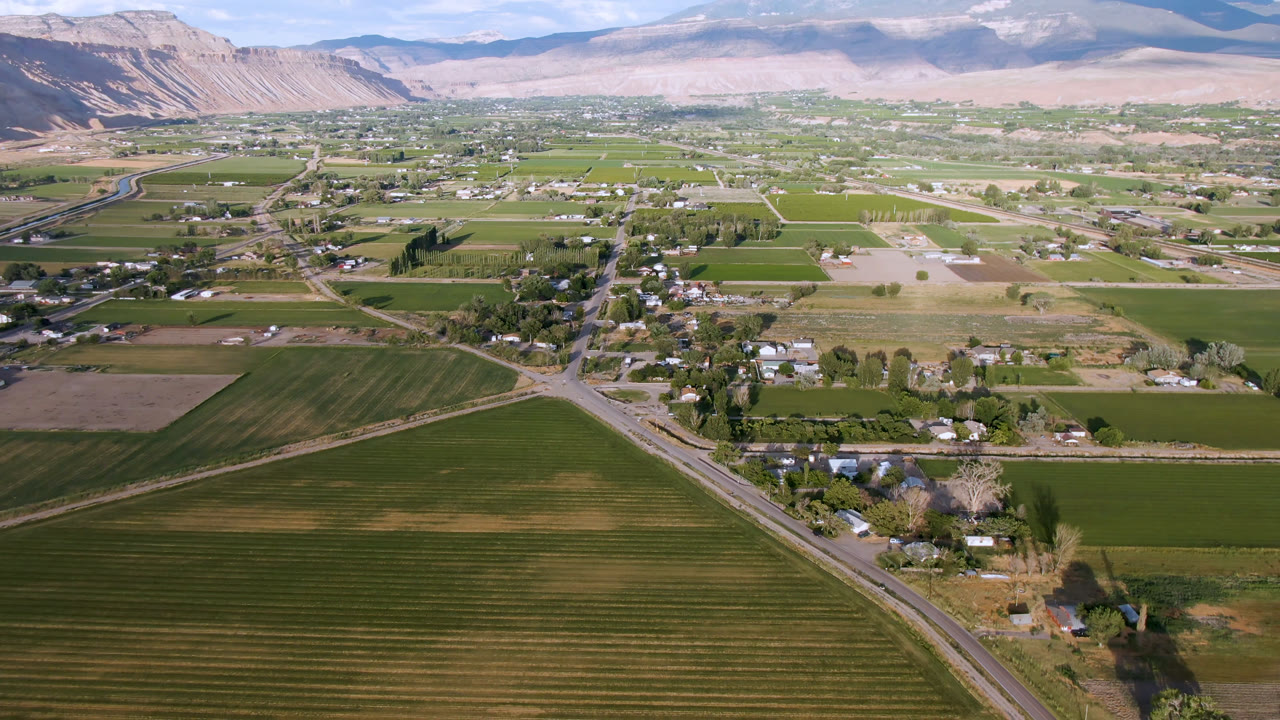 Opening aerial frame over Palisades, Colorado, showing green field rows, a diagonal road, scattered homes, trees, dry hills, distant mountains, and no visible people.