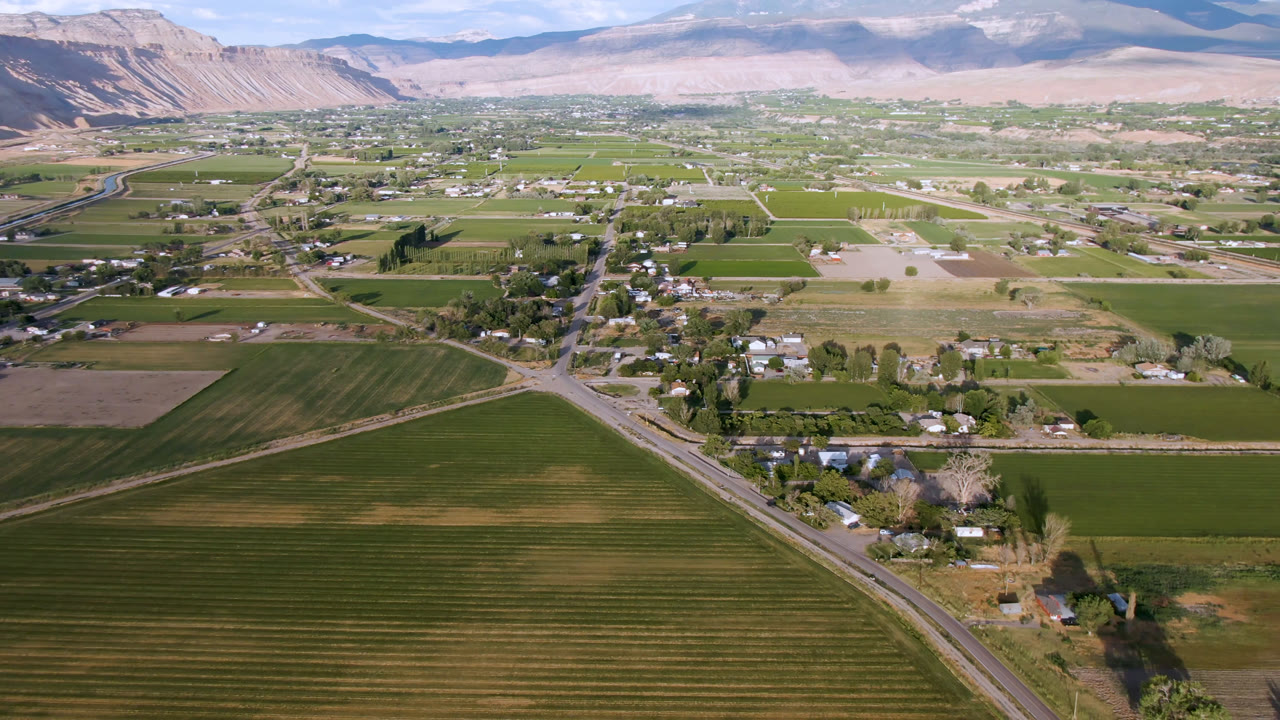 Second aerial frame over Palisades, Colorado, with a road crossing the lower left, rectangular farm plots, clustered homes, tree lines, pale hills, mountains, and no visible people.