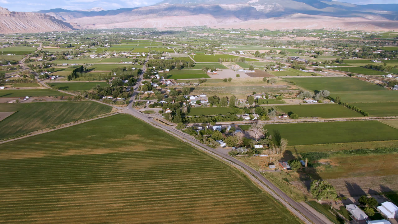 Second aerial frame over Palisades, Colorado showing a broad patchwork of fields, a road cutting through the lower frame, small rural structures, distant settlement, mountains, clouded sky, and no visible people.