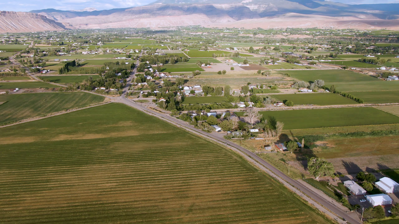 Drone-height view of patterned fields, a rural road, clustered trees, small buildings, and mountains.