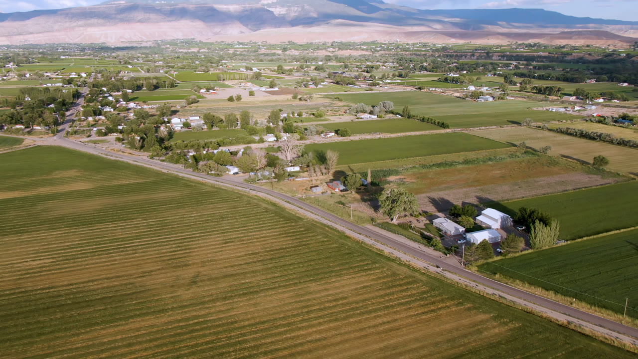 A wide aerial frame over Palisades, Colorado shows green and tan farm fields, a diagonal rural road, scattered buildings, trees, distant pale hills, and no visible people.