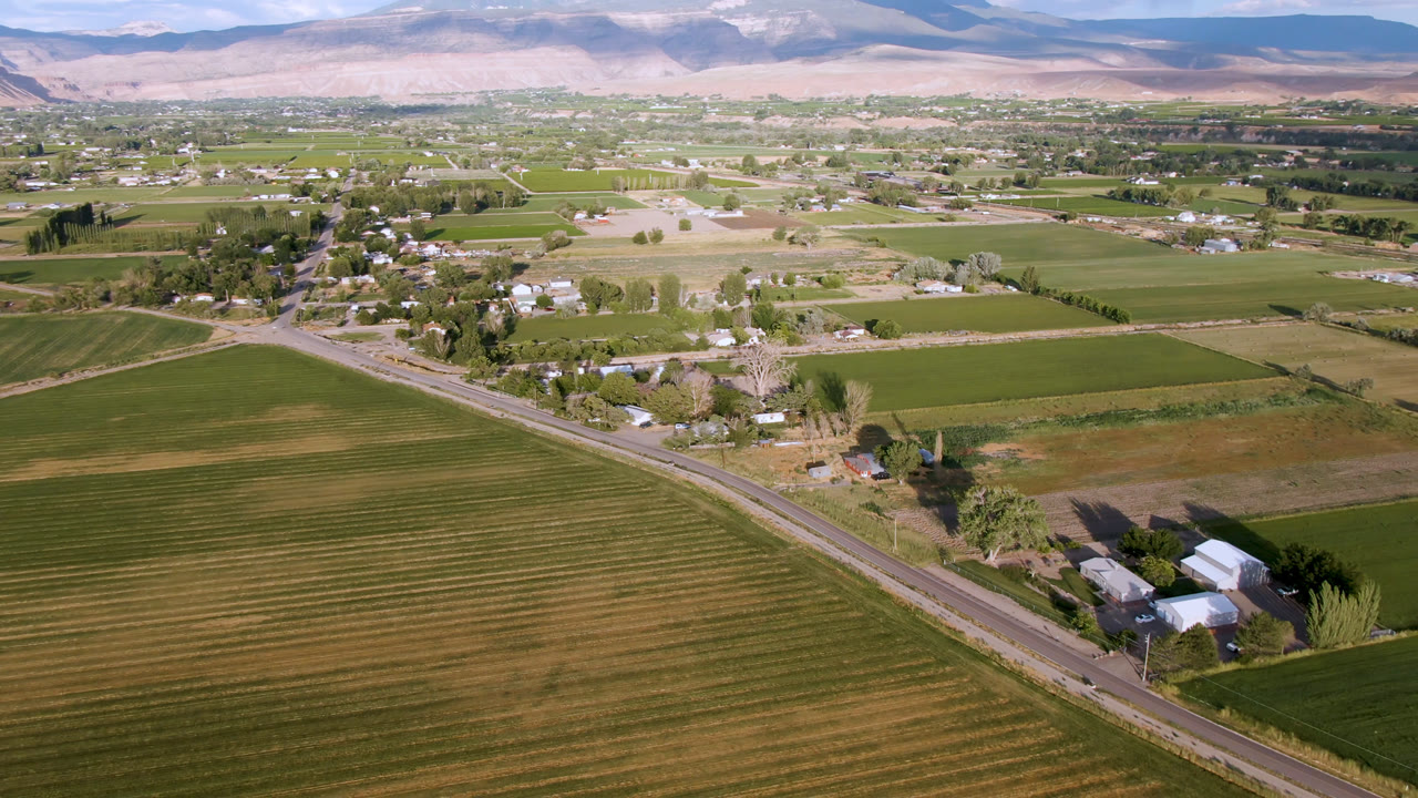 One second into the Palisades, Colorado aerial, furrowed fields fill the foreground while the road and rural homes sit below a mountain horizon with no visible people.