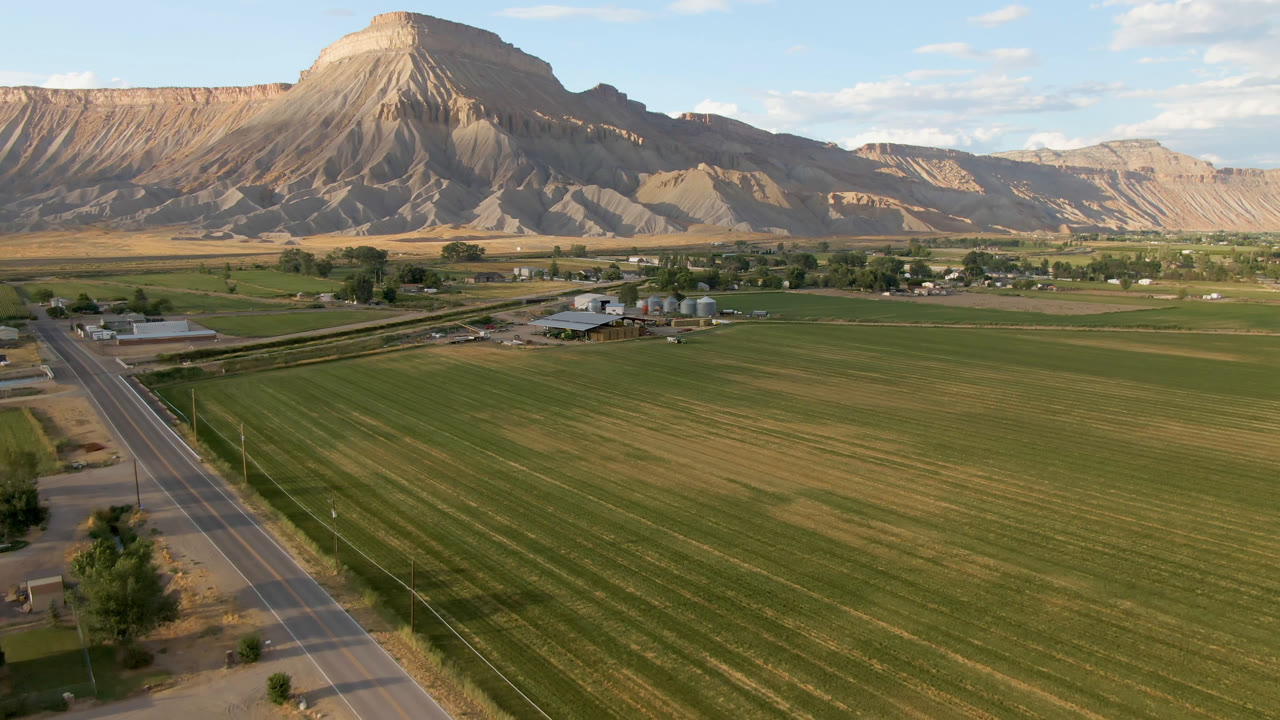 Aerial frame from Palisades, Colorado showing a paved road at left, broad green fields, low farm buildings, a tan mountain ridge, scattered clouds, and no visible people.
