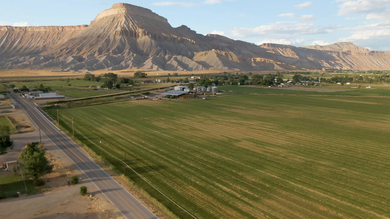 Drone-height view of fields, roadside trees, farm structures, and a flat-topped mountain.