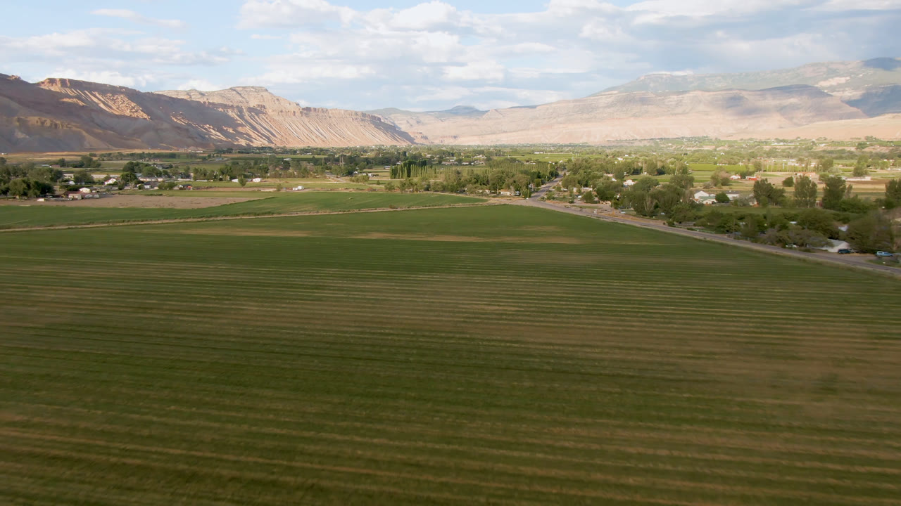 Wide aerial frame from Palisades, Colorado showing green field rows in the foreground, a road and small town beyond, pale mountains on the horizon, and no visible people.