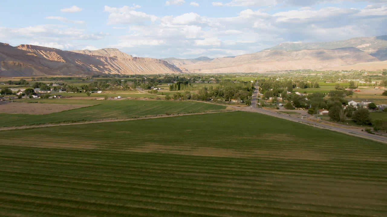 Early aerial frame over Palisades, Colorado, showing darker green field bands in front, a diagonal road near the center, low buildings among trees, and no visible people.