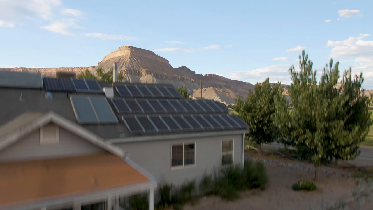 Elevated view in Palisades, Colorado showing a solar-roof house near trees, a fenced yard, distant fields, mesas, and no visible people.