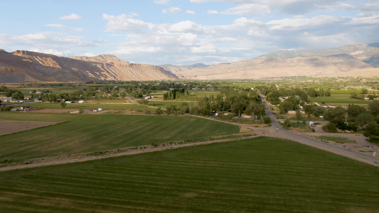 Opening wide aerial frame over Palisades, Colorado with green fields in the foreground, a road junction near the center, scattered buildings, tan mountains, clouds, and no visible people.