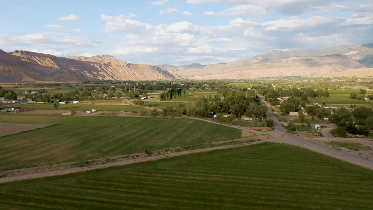 At 0:01, the Palisades, Colorado view holds on green farmland with a diagonal road, small buildings and trees in the midground, distant tan ridges, and no visible people.