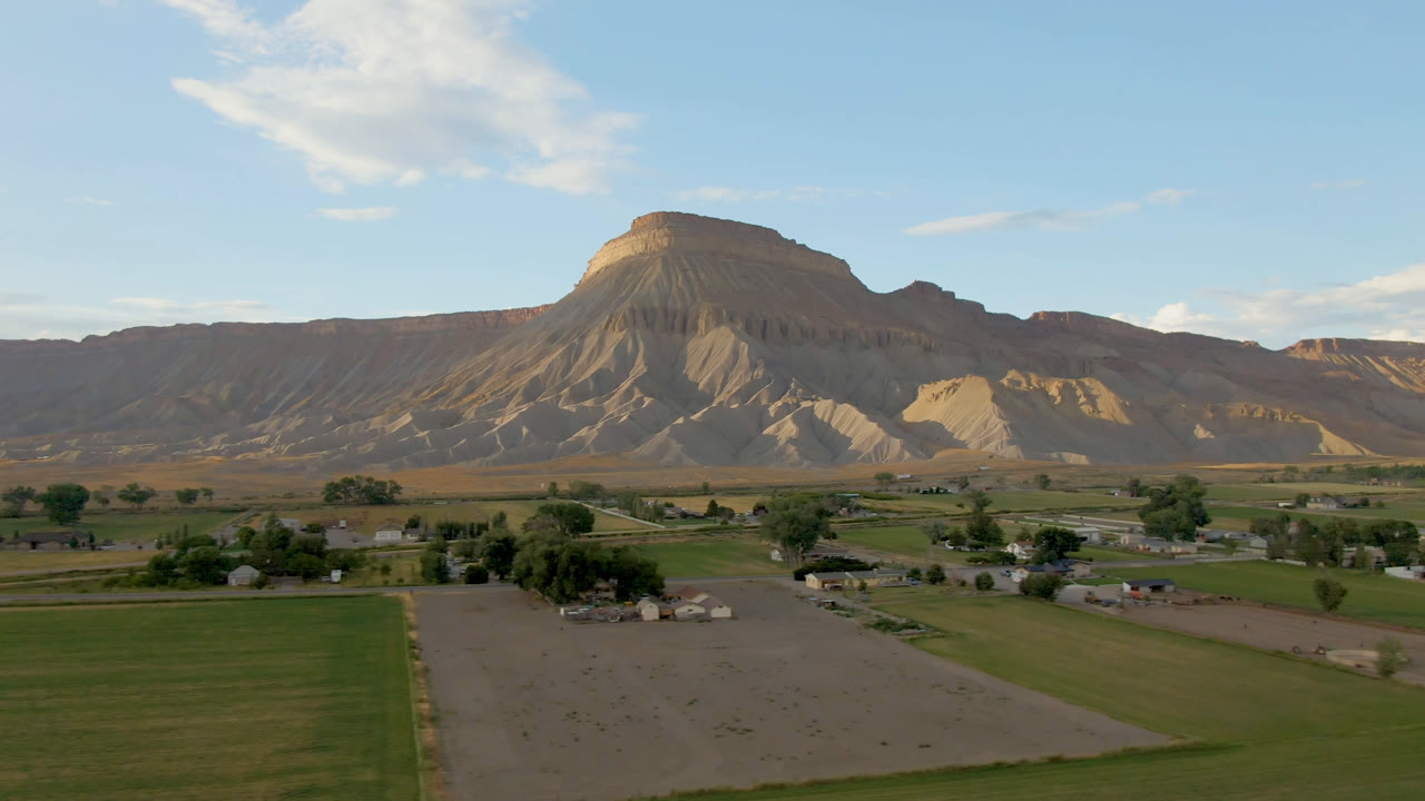 Wide aerial view of fields, small buildings, trees, and mesa cliffs under blue sky.