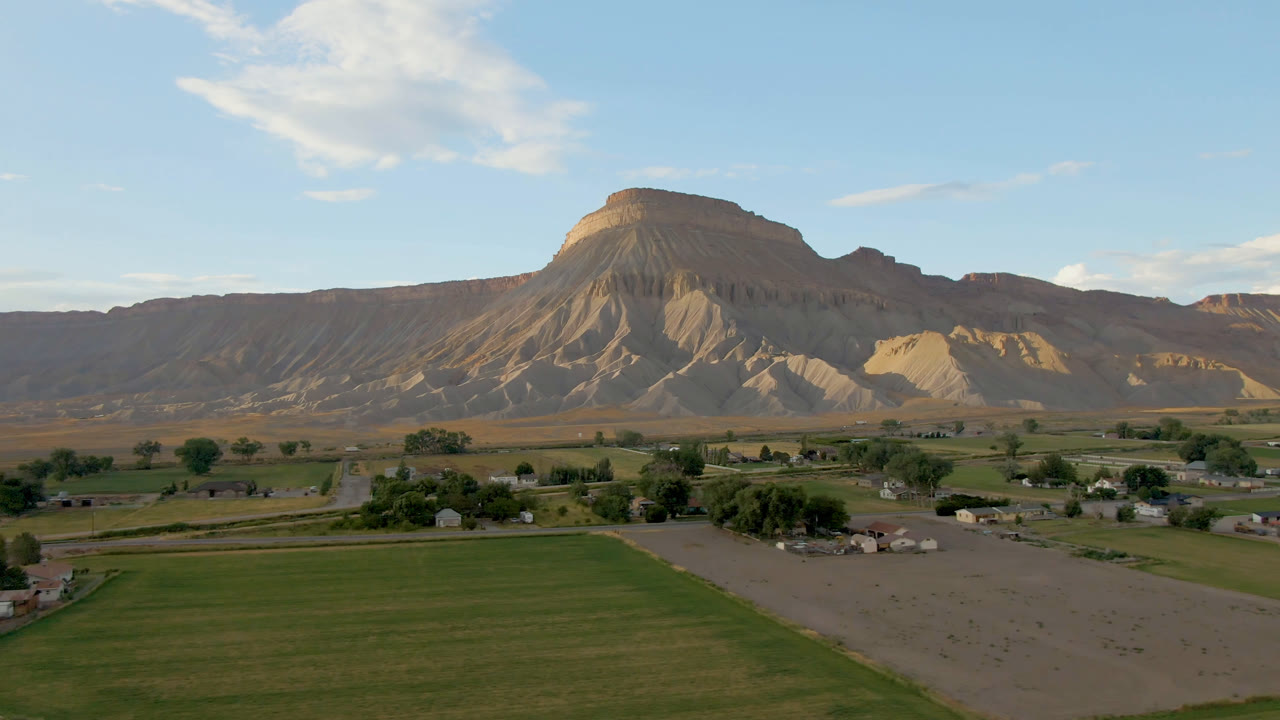 Wide rural landscape with green field, road, small buildings, and mesa ridge