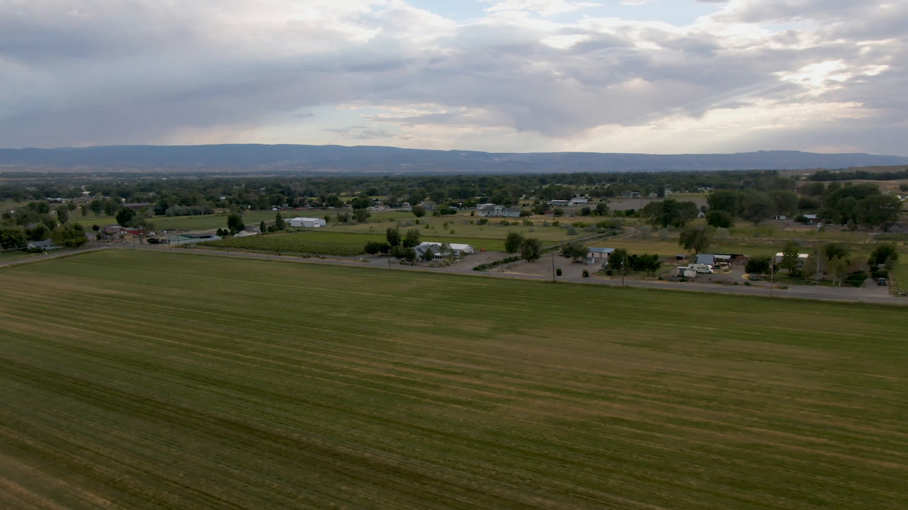 Wide aerial view over a green field, scattered houses, distant hills, and cloudy sky.