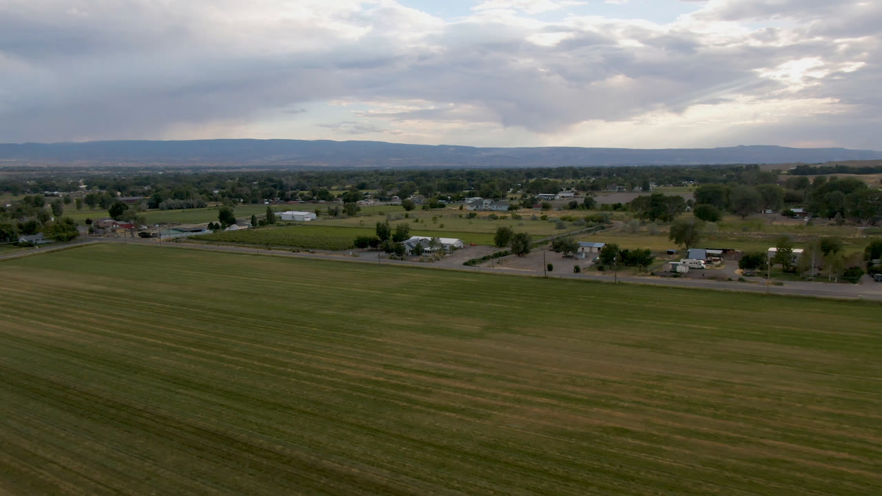 Second frame over Palisades, Colorado, showing more of the open field foreground, a thin road and clustered rural houses in the midground, distant hills, overcast sky, and no visible people.