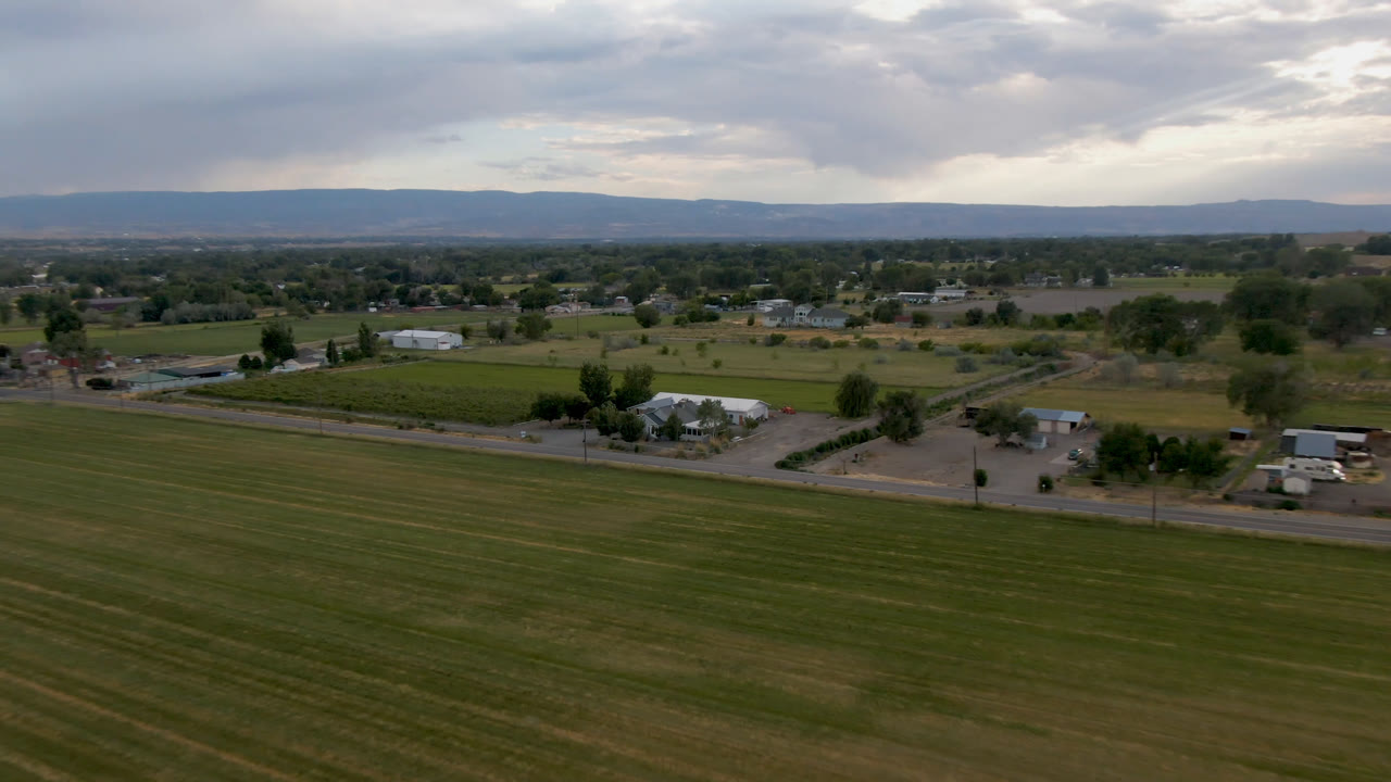 Wide aerial view of fields, a road, farm buildings, trees, and distant hills under clouds.