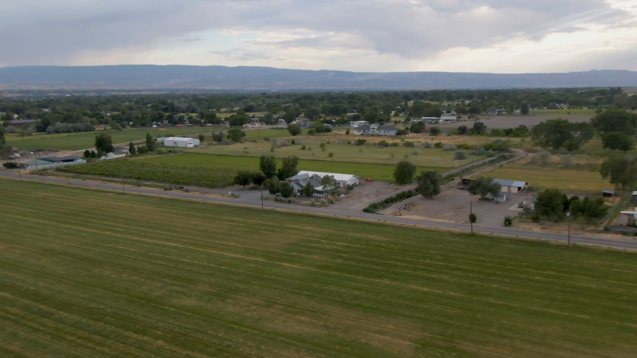 Second aerial frame over Palisades, Colorado showing striped fields, a straight road, clustered farm structures, tree rows, distant houses, low hills, and no visible people.