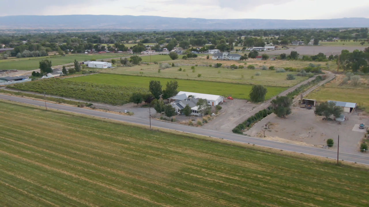 Aerial view of fields, a road, farm buildings, and distant hills under clouds.