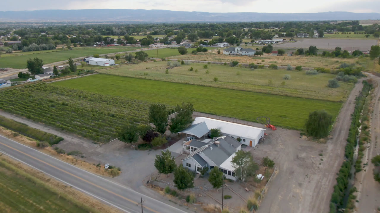 Drone-wide view in Palisades, Colorado with the road crossing the lower frame, a farmhouse and white outbuilding near trees, cultivated fields behind them, and no visible people.