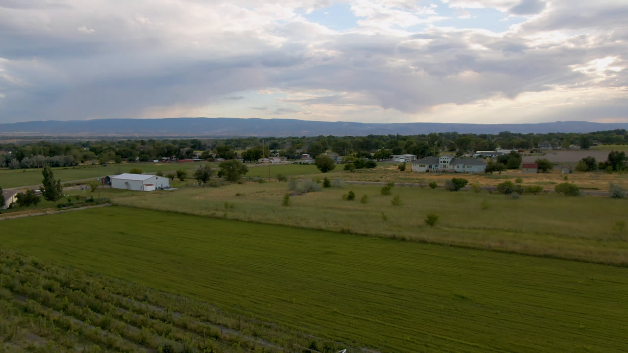 Aerial frame over a green Palisades, Colorado field with a pale farm building at left, scattered trees and homes in the distance, low hills on the horizon, and no visible people.