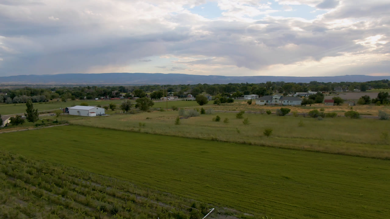 Aerial landscape frame from Palisades, Colorado with green field rows in front, small rural buildings beyond, a hazy hill line, heavy cloud cover, and no visible people.