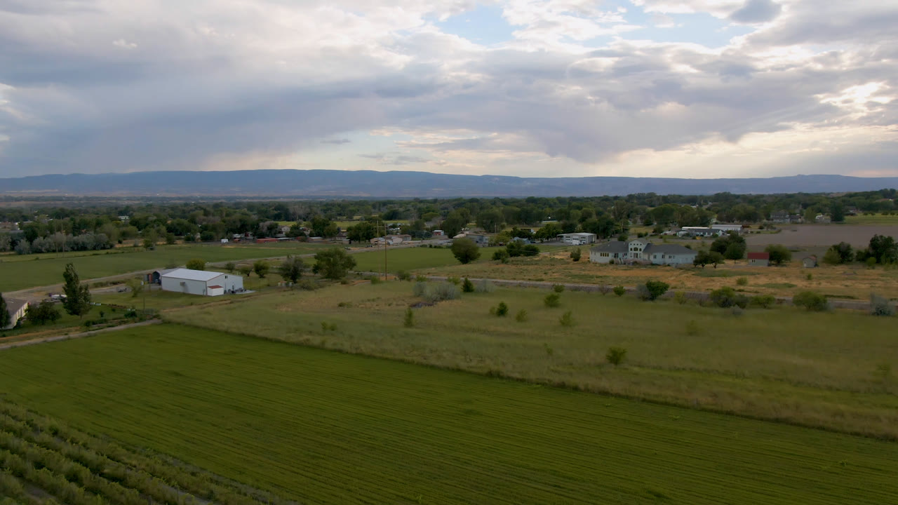 Wide aerial frame from Palisades, Colorado showing green agricultural fields in front, a white farm building near a road, scattered town structures, distant hills, and no visible people.