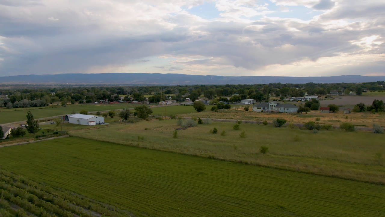 Palisades, Colorado aerial view with a broad green field in the foreground, the white farm building shifted slightly left of center, a thin road, distant houses, hills, and no visible people.