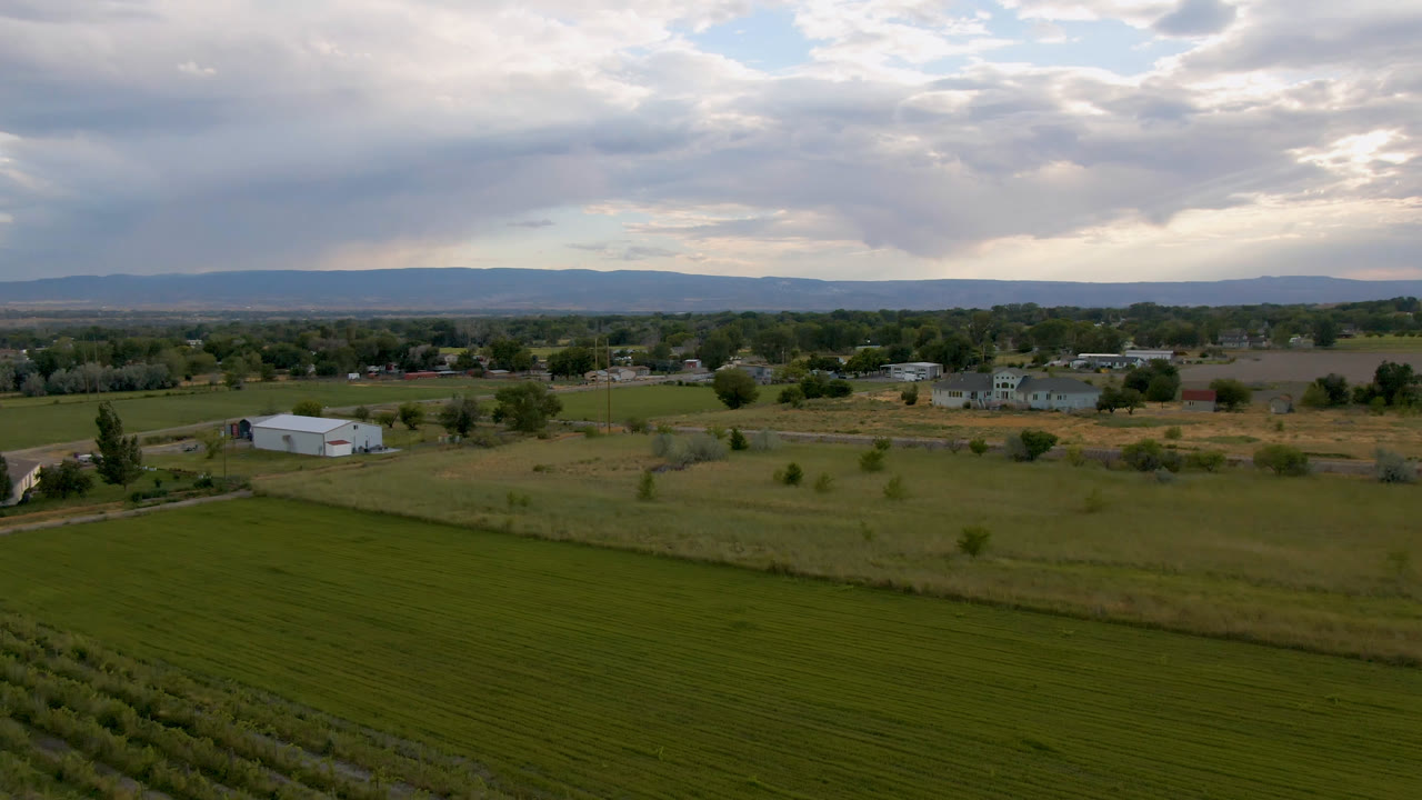 Drone-wide rural landscape with field rows, white outbuilding, scattered homes, trees, hills, and cloud layers.