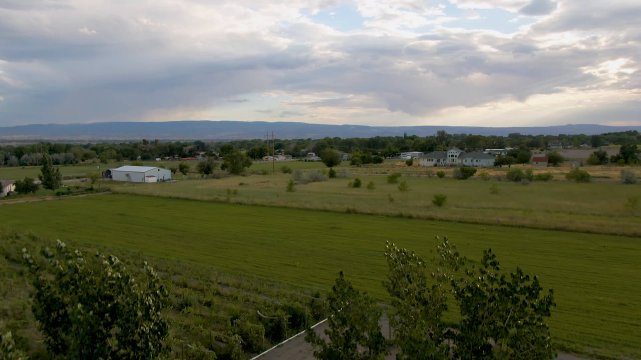 Wide Palisades, Colorado frame showing a mowed green field, rural homes, scattered trees, distant hills, and a bright break in the cloudy sky, with no visible people.