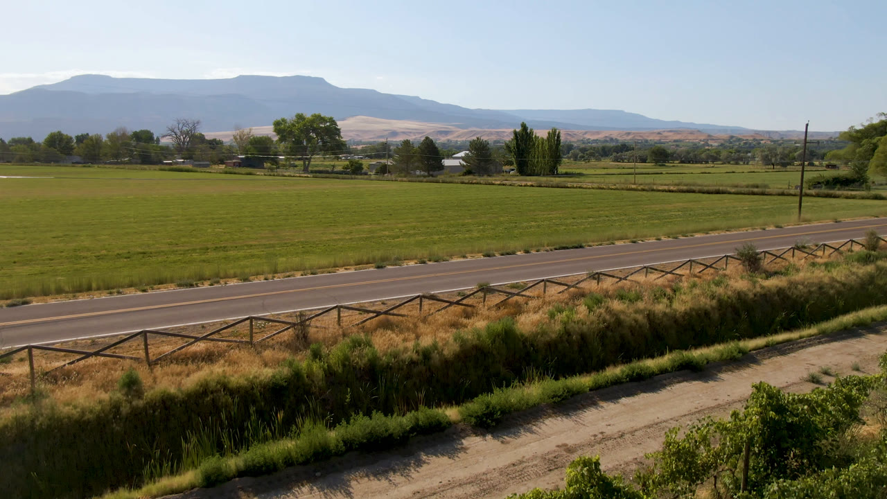 Elevated view in Palisades, Colorado of a paved road, roadside fence, green fields, trees, and distant mountains under a clear sky, with no visible people.