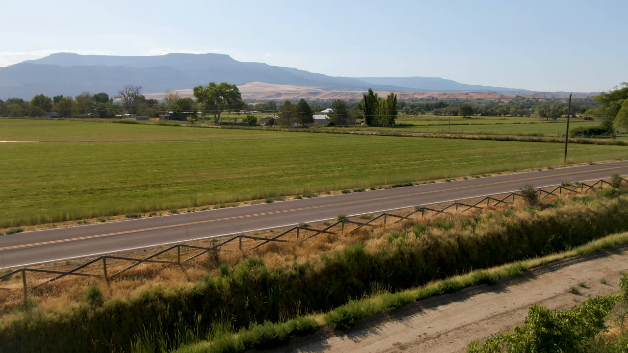 Wide Palisades, Colorado frame with a two-lane road crossing the foreground, a fence line, green farm fields, scattered trees, and a hazy mountain ridge, with no visible people.