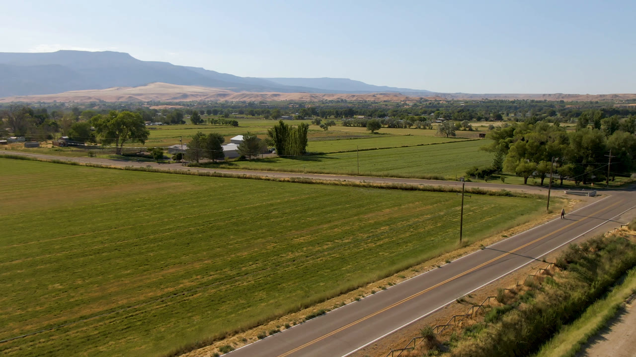 At 0:01, the Palisades, Colorado landscape remains wide, with a road junction in the foreground, rectangular green fields, tree rows, small farm structures, and a hazy mountain ridge in the distance.