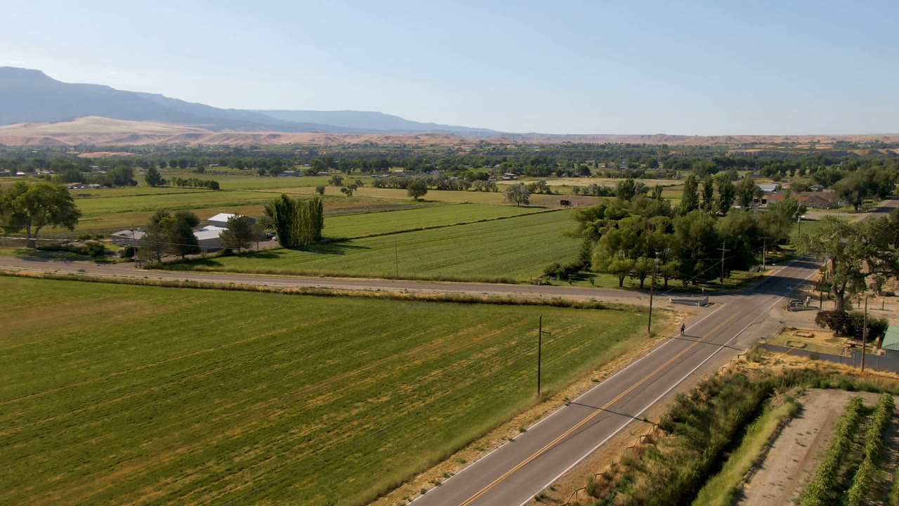 Aerial view in Palisades, Colorado, with the road slightly more centered below the fields, farm buildings among trees, distant hills, and no visible people.